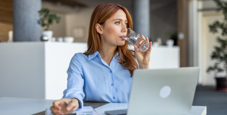 A professional businesswoman takes a short break at her desk in a modern office environment. She enjoys a refreshing sip of water while working on her laptop, balancing productivity with self-care.