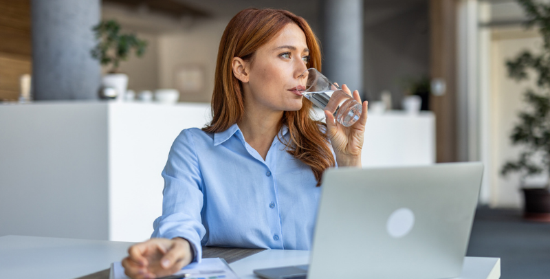 A professional businesswoman takes a short break at her desk in a modern office environment. She enjoys a refreshing sip of water while working on her laptop, balancing productivity with self-care.